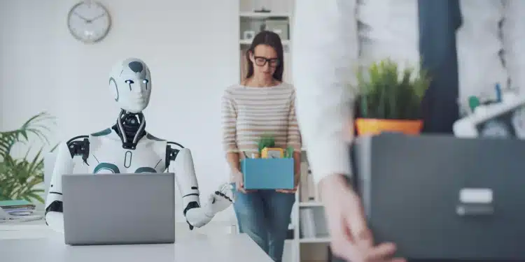 Robô sentado em frente a notebook enquanto pessoas carregam caixas, deixando o escritório.