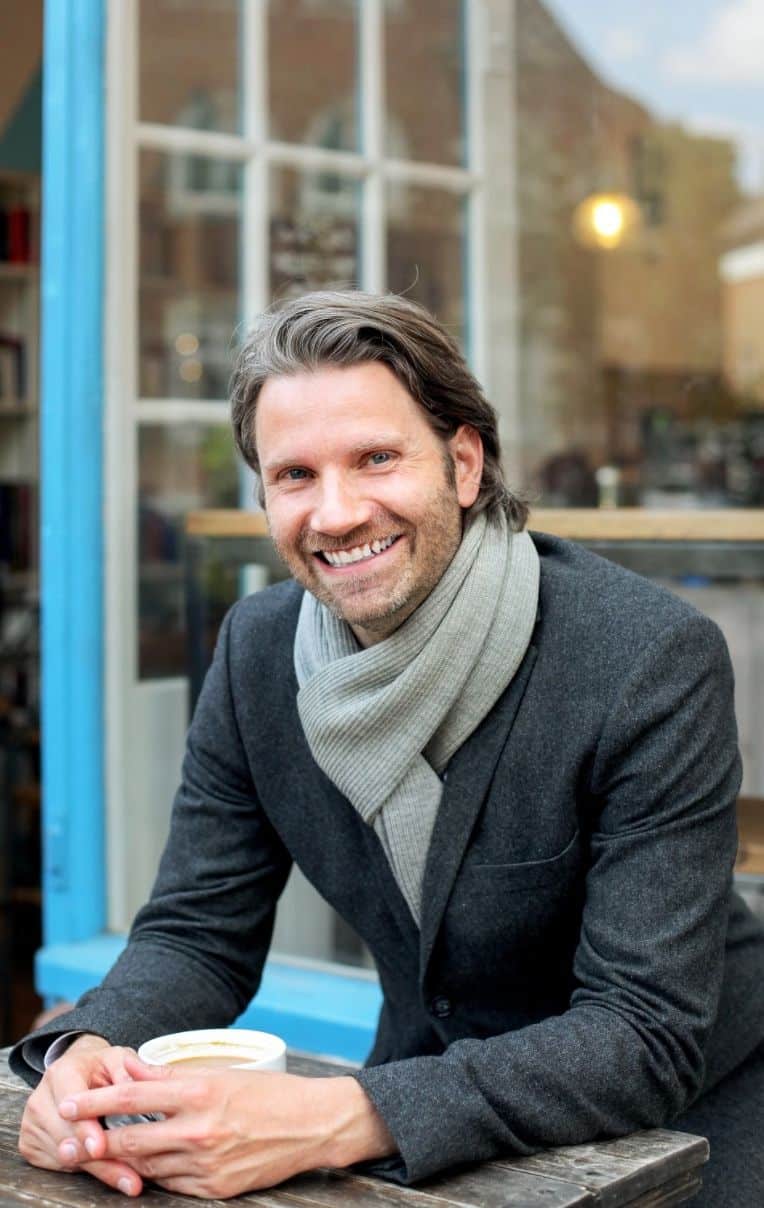 Homem sorrindo sentado à mesa com xícara de café e cachecol cinza.