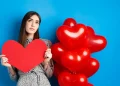 Mulher segurando coração de papel vermelho, ao lado de balões vermelhos em forma de coração, simbolizando o amor como chave para a felicidade.