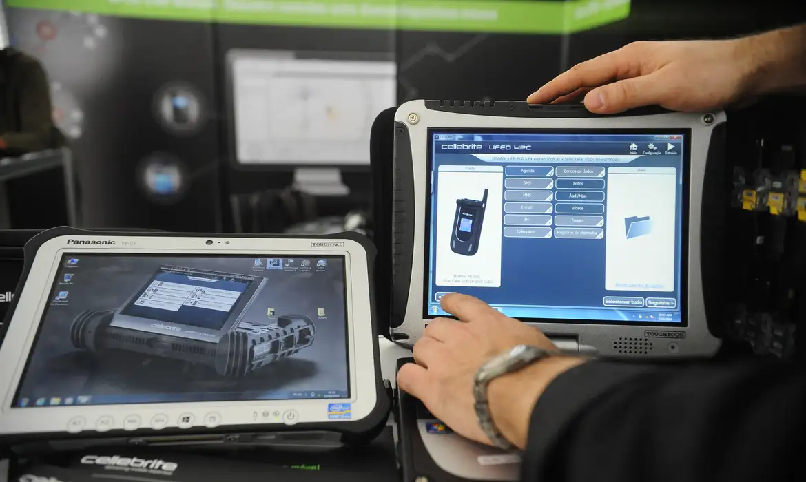 Mãos manipulando tablets robustos com software Cellebrite de extração de dados exibido na tela