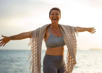 Mulher madura de cabelos curtos sorrindo com braços abertos na praia durante o pôr do sol, usando roupas esportivas e uma echarpe.