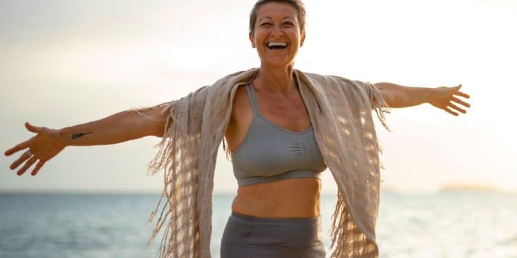 Mulher madura de cabelos curtos sorrindo com braços abertos na praia durante o pôr do sol, usando roupas esportivas e uma echarpe.