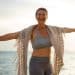 Mulher madura de cabelos curtos sorrindo com braços abertos na praia durante o pôr do sol, usando roupas esportivas e uma echarpe.
