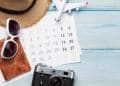 Visão aérea de um calendário sobre mesa de madeira azul, cercado por óculos de sol brancos, chapéu de palha, câmera fotográfica retrô e um avião de brinquedo em miniatura.