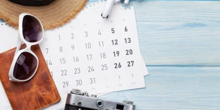 Visão aérea de um calendário sobre mesa de madeira azul, cercado por óculos de sol brancos, chapéu de palha, câmera fotográfica retrô e um avião de brinquedo em miniatura.