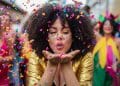 Mulher jovem de cabelos cacheados e jaqueta dourada soprando confetes coloridos em uma festa de rua animada durante o Carnaval.
