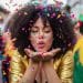 Mulher jovem de cabelos cacheados e jaqueta dourada soprando confetes coloridos em uma festa de rua animada durante o Carnaval.