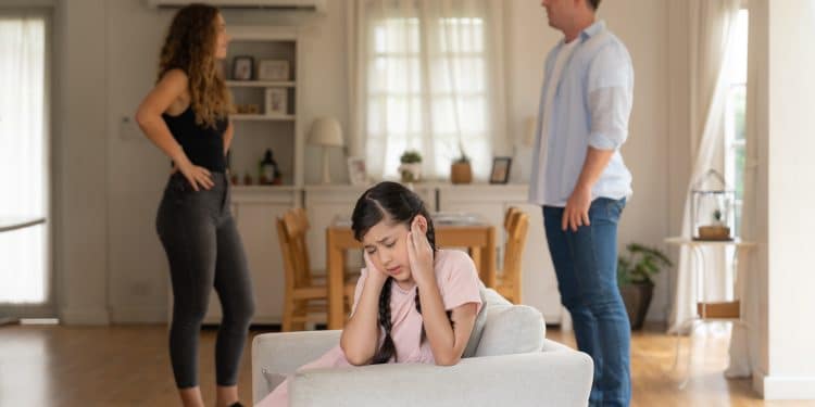 Criança sentada no sofá com as mãos nas orelhas enquanto seus pais discutem ao fundo, demonstrando tensão familiar.
