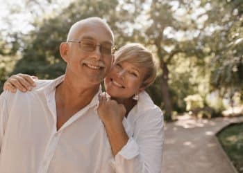 Casal maduro sorridente, com a mulher encostando o rosto no ombro do homem, em um parque iluminado pelo sol, ilustrando a felicidade e a cumplicidade na maturidade.