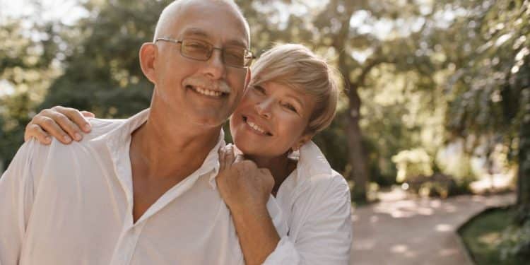 Casal maduro sorridente, com a mulher encostando o rosto no ombro do homem, em um parque iluminado pelo sol, ilustrando a felicidade e a cumplicidade na maturidade.