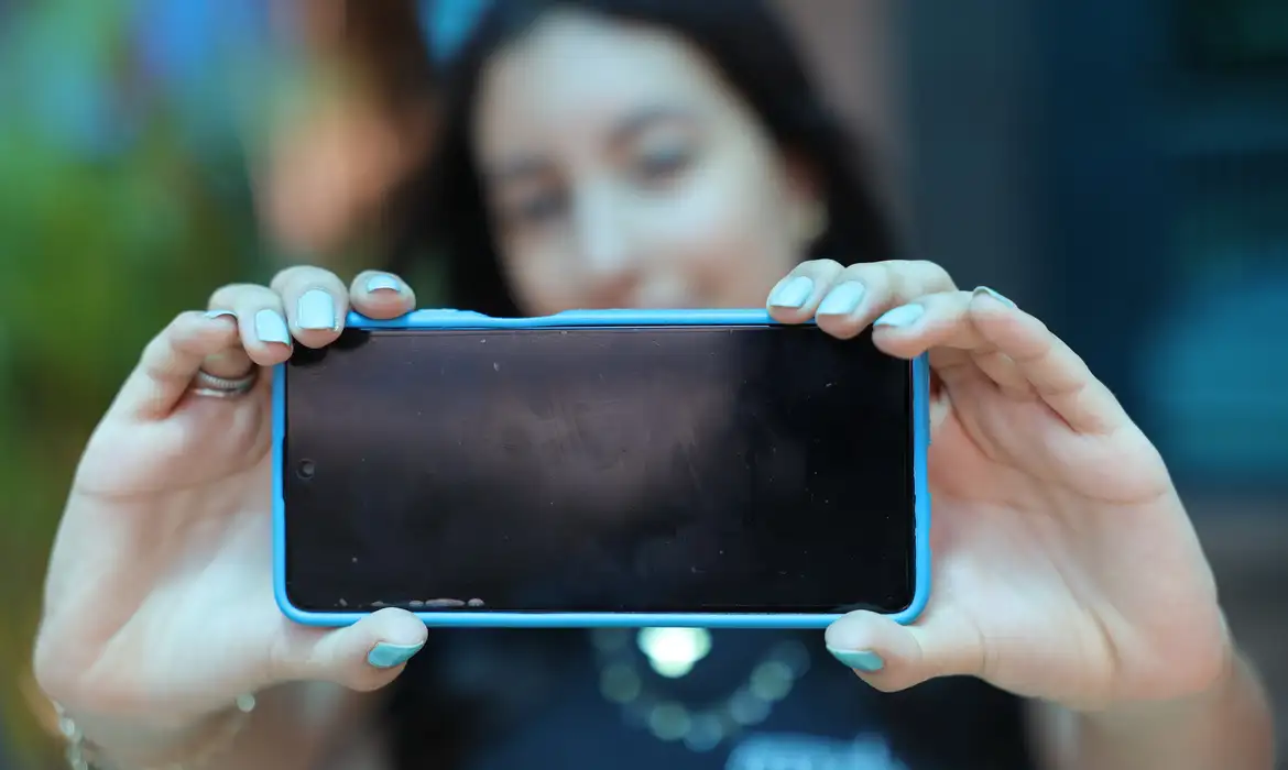 Mulher jovem com o rosto desfocado ao fundo, segurando um smartphone na horizontal com ambas as mãos em destaque. O aparelho possui uma capa azul claro e a tela está voltada para a câmera, simbolizando a posse segura e o controle do dispositivo.