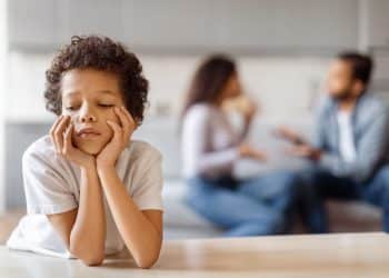 Criança com expressão triste, descansando o rosto nas mãos enquanto observa um ambiente familiar tenso.