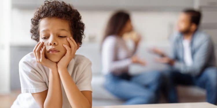 Criança com expressão triste, descansando o rosto nas mãos enquanto observa um ambiente familiar tenso.