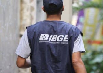 Homem de costas usando o colete azul oficial do IBGE, com o logotipo do Instituto Brasileiro de Geografia e Estatística em destaque, caminhando por uma rua estreita durante o trabalho de coleta de dados.