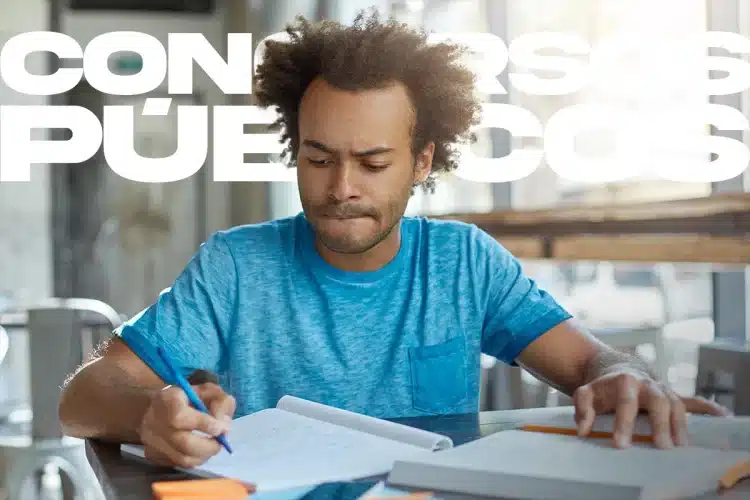 Homem com camiseta azul estudando em mesa com cadernos e caneta em ambiente iluminado.