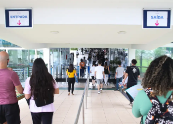 Candidatos entrando em local de prova de concurso público com placas de entrada e saída