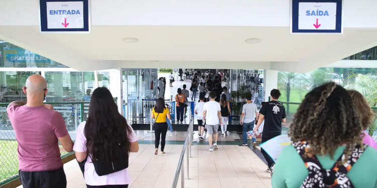 Candidatos entrando em local de prova de concurso público com placas de entrada e saída