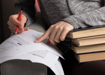 Candidata estudando com lápis vermelho apontando para documentos ao lado de pilha de livros em preparação para o concurso SAMAE Campos Novos SC