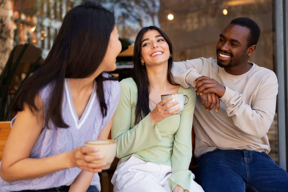Três amigos — uma mulher asiática, uma mulher latina e um homem negro — sentados em um banco de madeira ao ar livre, sorrindo e conversando enquanto seguram xícaras de café.