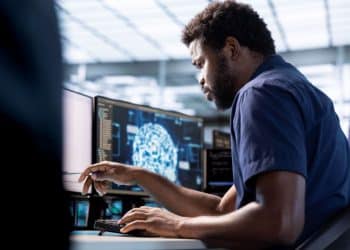 Homem negro trabalhando concentrado em um escritório moderno, operando dois monitores que exibem códigos de programação e uma representação gráfica de rede neural de Inteligência Artificial.