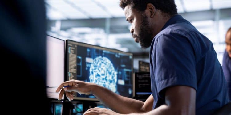 Homem negro trabalhando concentrado em um escritório moderno, operando dois monitores que exibem códigos de programação e uma representação gráfica de rede neural de Inteligência Artificial.
