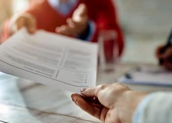 Uma pessoa entrega um currículo para um recrutador durante uma entrevista de emprego em uma mesa de madeira clara, com foco na troca de documentos.