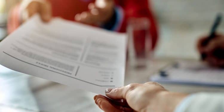 Uma pessoa entrega um currículo para um recrutador durante uma entrevista de emprego em uma mesa de madeira clara, com foco na troca de documentos.