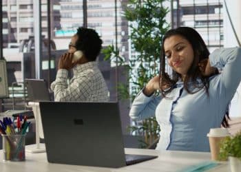 Mulher em um escritório moderno alongando o pescoço e os ombros com expressão de fadiga enquanto está sentada à frente de um laptop.