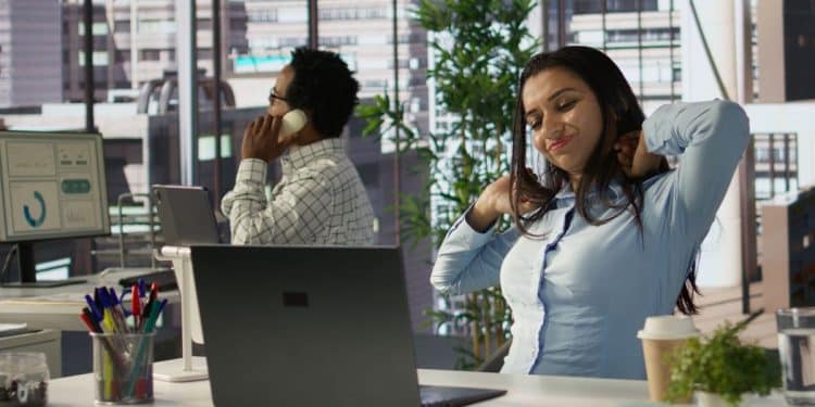 Mulher em um escritório moderno alongando o pescoço e os ombros com expressão de fadiga enquanto está sentada à frente de um laptop.