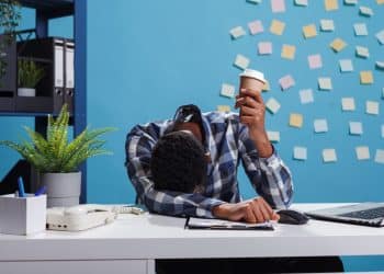 Um trabalhador de pele escura com cabelo crespo, vestindo camisa xadrez azul e branca, está com a cabeça abaixada sobre a mesa em um escritório moderno de fundo azul com post-its. Ele segura um copo de café descartável, visivelmente exausto ao lado de um notebook e um telefone fixo.