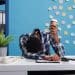 Um trabalhador de pele escura com cabelo crespo, vestindo camisa xadrez azul e branca, está com a cabeça abaixada sobre a mesa em um escritório moderno de fundo azul com post-its. Ele segura um copo de café descartável, visivelmente exausto ao lado de um notebook e um telefone fixo.