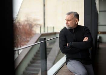 Close-up de um homem maduro com barba grisalha e blusa preta, sentado de braços cruzados no deck de uma sacada, olhando para o horizonte com expressão pensativa.