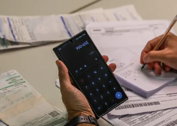 Mão segura celular com calculadora e outra faz anotações entre boletos