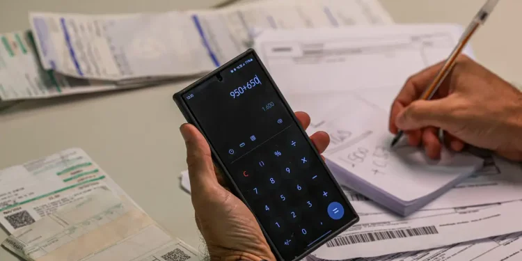 Mão segura celular com calculadora e outra faz anotações entre boletos