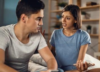 Homem e mulher sentados em um quarto conversam de forma séria, expressando emoções durante um conflito no relacionamento.