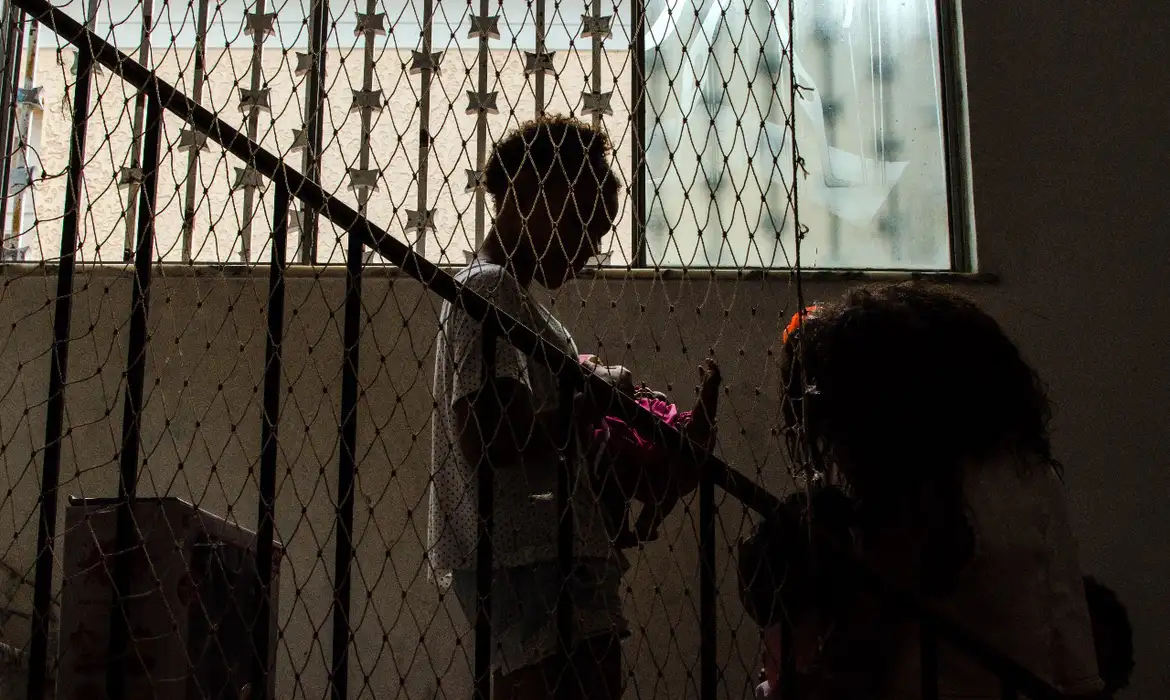 Duas crianças interagem próximas a uma janela, separadas por tela de proteção em escada interna