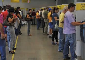 Interior de uma agência bancária com uma longa fila de pessoas aguardando atendimento. À direita, terminais de autoatendimento (caixas eletrônicos) cinzas com sinalização amarela escrita "Saques, Extratos, Pagamentos, Transferências".