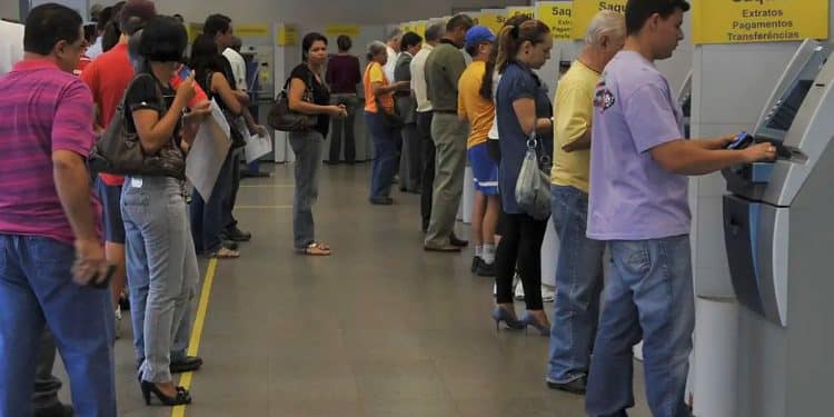 Interior de uma agência bancária com uma longa fila de pessoas aguardando atendimento. À direita, terminais de autoatendimento (caixas eletrônicos) cinzas com sinalização amarela escrita "Saques, Extratos, Pagamentos, Transferências".