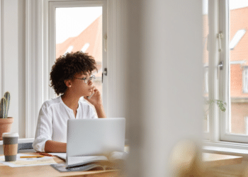 Uma mulher jovem com cabelo cacheado curto e óculos de grau está sentada à mesa de trabalho em frente a uma janela iluminada. Ela veste uma blusa branca, segura um smartphone no ouvido em uma chamada e olha pensativa para o lado. Sobre a mesa de madeira, há um laptop prateado, um copo de café para viagem, papéis e um pequeno cacto em um vaso de barro