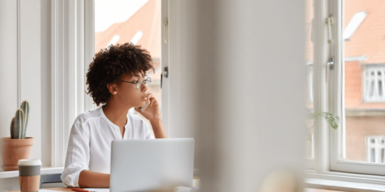 Uma mulher jovem com cabelo cacheado curto e óculos de grau está sentada à mesa de trabalho em frente a uma janela iluminada. Ela veste uma blusa branca, segura um smartphone no ouvido em uma chamada e olha pensativa para o lado. Sobre a mesa de madeira, há um laptop prateado, um copo de café para viagem, papéis e um pequeno cacto em um vaso de barro