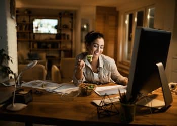 Mulher sorrindo enquanto come salada e trabalha em computador de mesa durante expediente noturno em home office