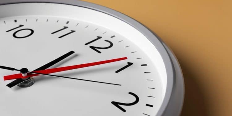 Close-up lateral de um relógio de parede redondo e branco sobre uma superfície de fundo ocre. O ponteiro de segundos vermelho está posicionado próximo ao número um, enquanto os ponteiros pretos de horas e minutos indicam aproximadamente dez horas e sete minutos. A iluminação suave destaca a curvatura da moldura cinza do relógio.