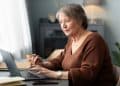 Mulher sênior de cabelos curtos grisalhos, vestindo uma blusa marrom, sentada à mesa e utilizando um notebook com foco e atenção em um ambiente doméstico iluminado.
