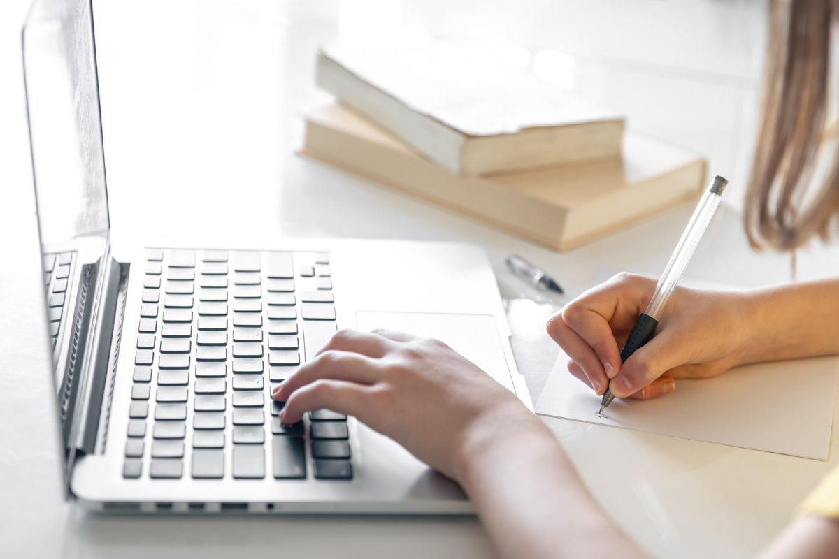 Pessoa escrevendo em uma folha enquanto usa um laptop para inscrição em concurso.