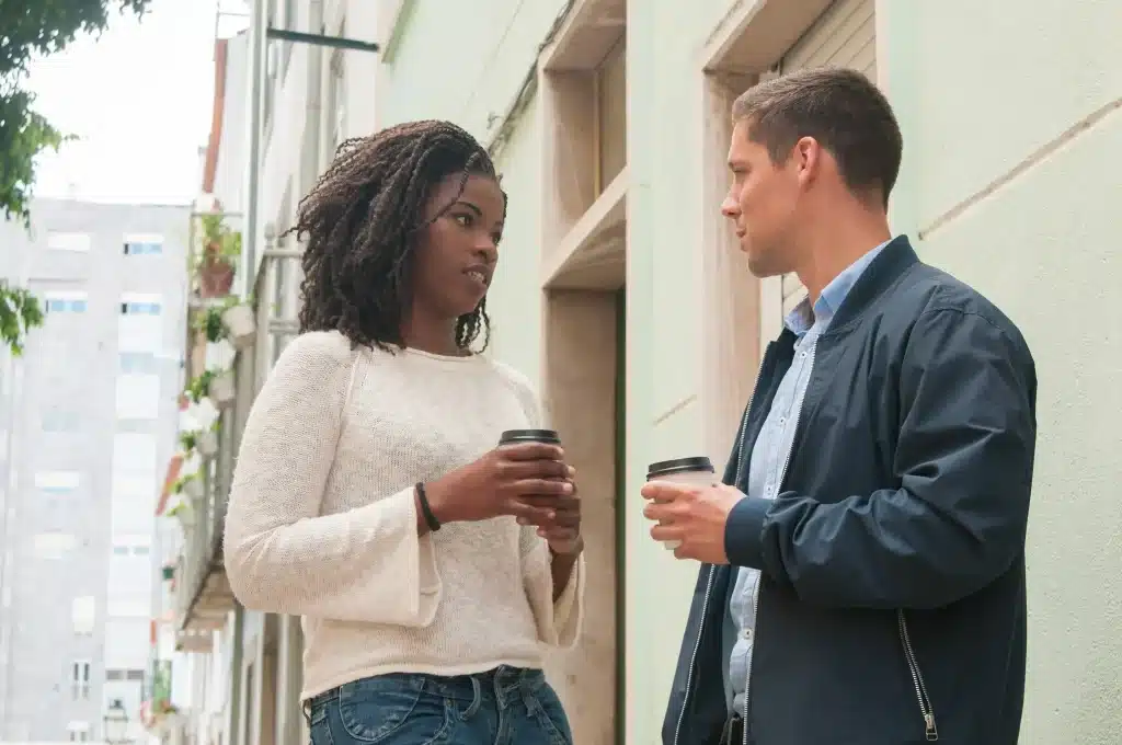 Homem e mulher conversando seriamente na rua enquanto seguram copos de café, representando diálogo assertivo e inteligência emocional.
