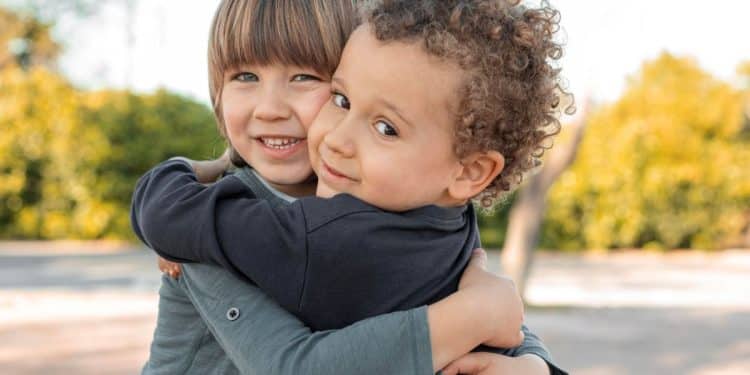 Duas crianças pequenas, um menino de cabelos loiros e um menino negro de cabelos cacheados, sorrindo e se abraçando carinhosamente em um fundo branco e limpo.
