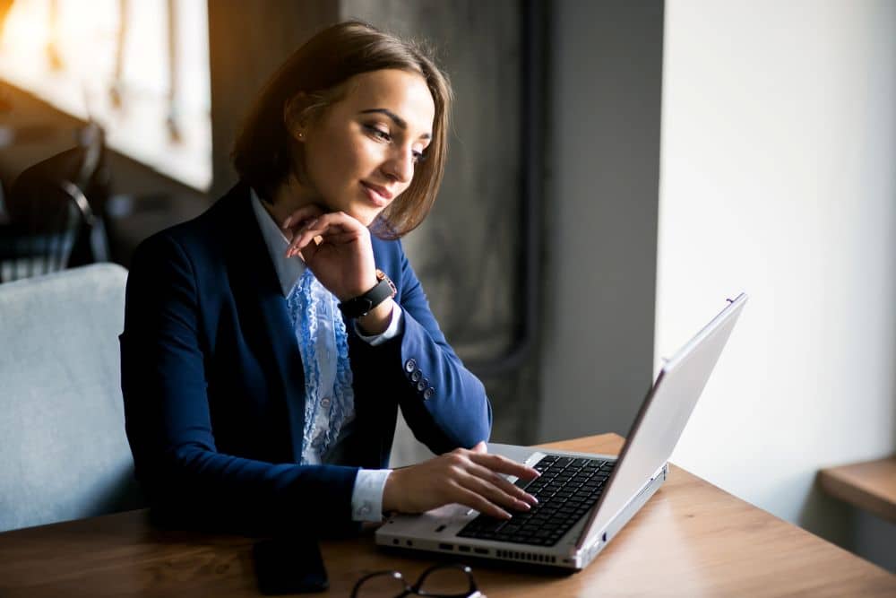 Mulher jovem de blazer azul marinho e camisa branca com detalhes em babados, sentada à mesa utilizando um notebook cinza. Ela tem o queixo apoiado na mão esquerda e demonstra expressão de foco ao olhar para a tela.