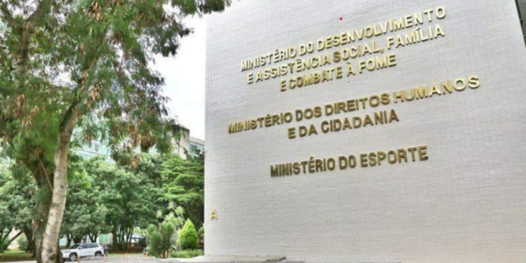 Vista lateral da fachada de um edifício governamental em Brasília, com paredes brancas e letras douradas que identificam o Ministério do Desenvolvimento Social, Família e Combate à Fome, o Ministério dos Direitos Humanos e o Ministério do Esporte. À esquerda, árvores verdes e um carro estacionado compõem o cenário sob luz natural.