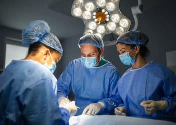 Três cirurgiões, dois homens e uma mulher, realizam um procedimento médico em um centro cirúrgico iluminado. Eles vestem uniformes azuis, máscaras, toucas e luvas descartáveis, focados no trabalho sob a luz de um foco cirúrgico de LED no teto.