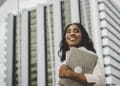 Mulher negra jovem e sorridente, vestindo camisa branca, segurando um notebook fechado contra o peito e olhando para o horizonte. Ao fundo, prédios comerciais modernos em tons de cinza e branco sob um céu claro.
